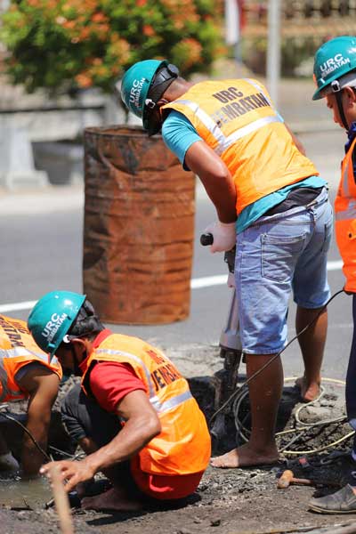 Construction workers performing a plate bearing test on a road, using tools and a jackhammer near a water-filled trench.
