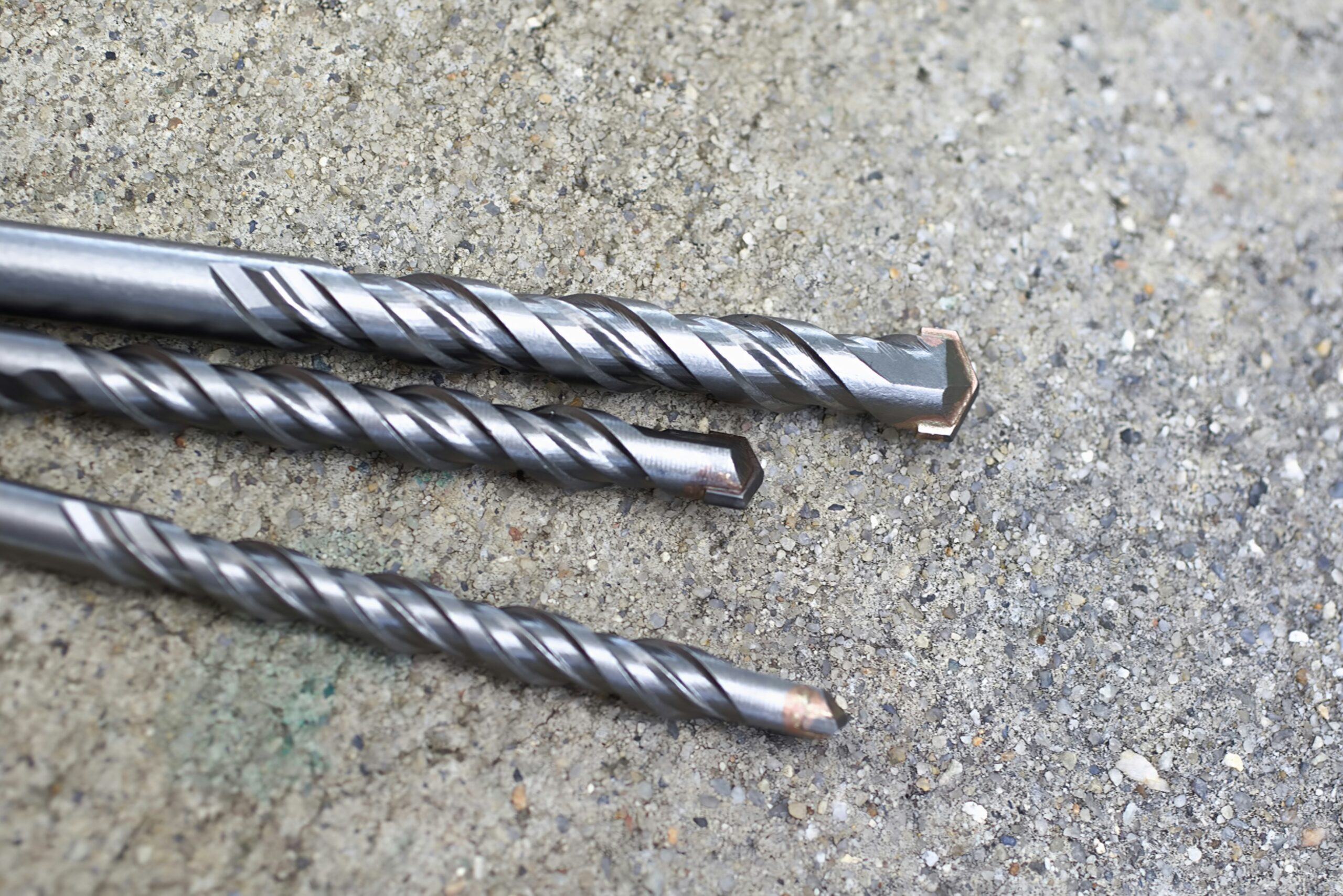 Close-up of three precision concrete drilling bits with carbide-tipped edges, arranged diagonally on a textured concrete surface, showcasing their helical design and durability for drilling into hard materials.